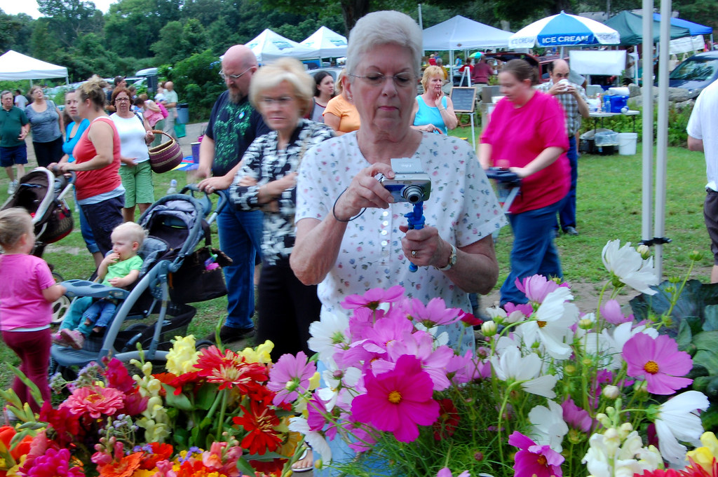 n old woman takes a picture of a florist’s booth at the Coventry Farmers Market